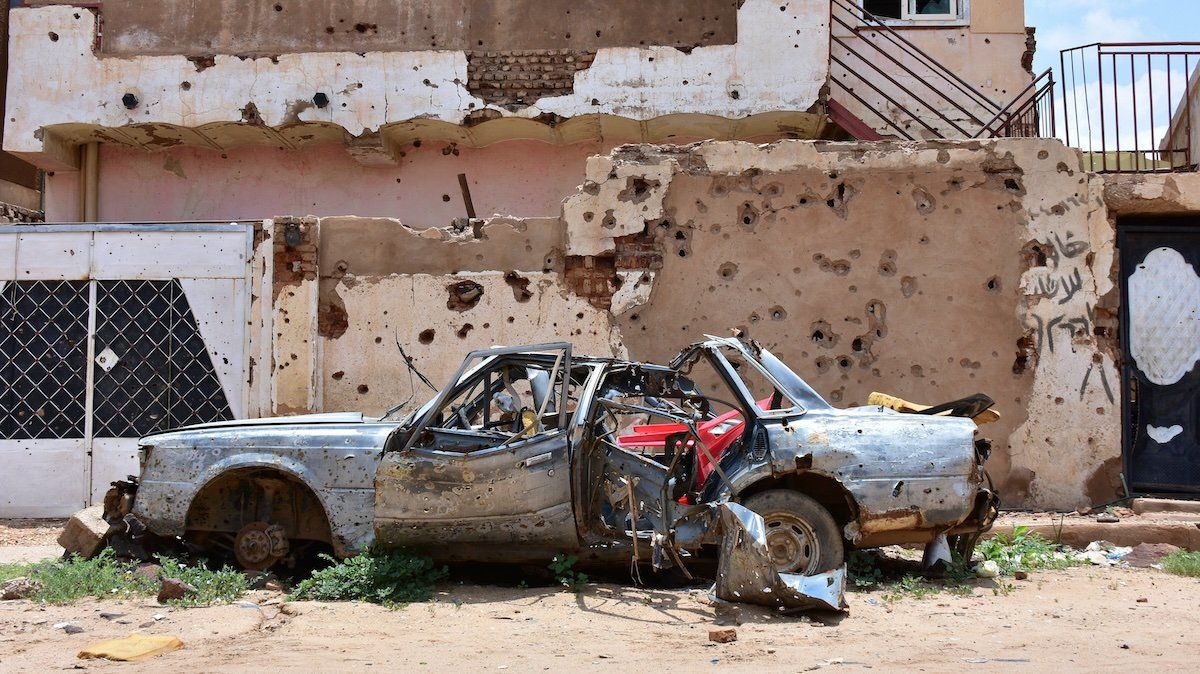 A destroyed car stands in front of a house full of bullet holes in Sudan.