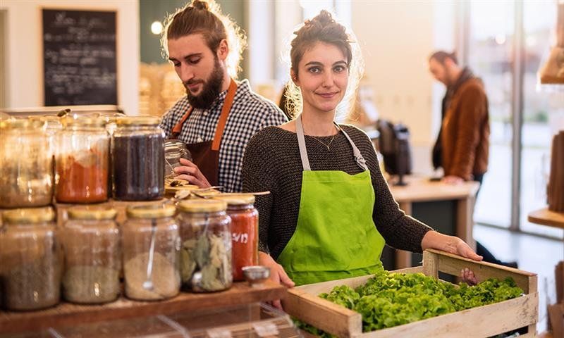 A woman holding a crate of leafy greens and a man behind her taking out spices from a jar