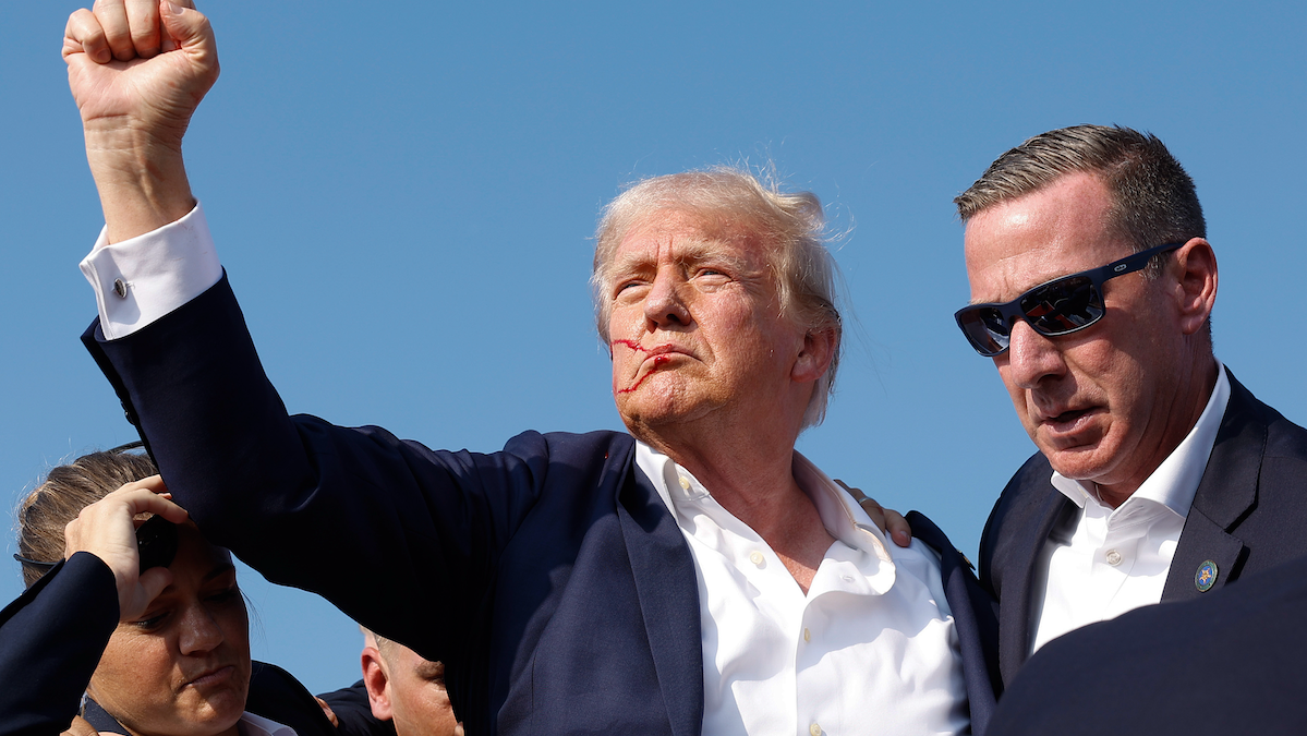 ​Former President Donald Trump, with his face bloodied by a shot that hit his right ear, raises his fist as he's rushed from a rally stage in Butler, PA.